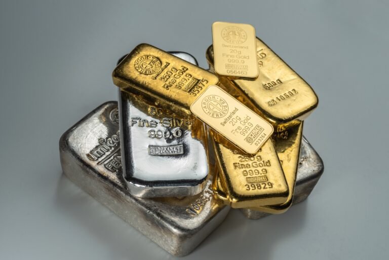 A pile of gold and silver bars of various weights from different manufacturers lies on a grey background.